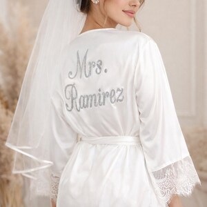May include: White satin bridal robe with lace trim and the name "Mrs. Ramirez" in silver glitter lettering on the back. The robe has a matching belt and is paired with a sheer veil.
