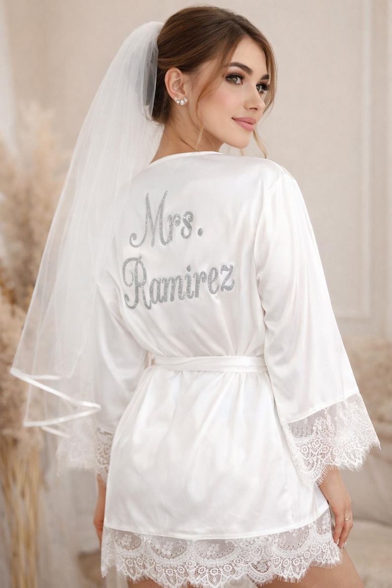 May include: White satin bridal robe with lace trim and the name "Mrs. Ramirez" in silver glitter lettering on the back. The robe has a matching belt and is paired with a sheer veil.