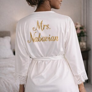 May include: White satin robe with lace trim and a matching belt. The back of the robe features the gold glitter text "Mrs. Nabavian". The robe has long sleeves and is knee-length.