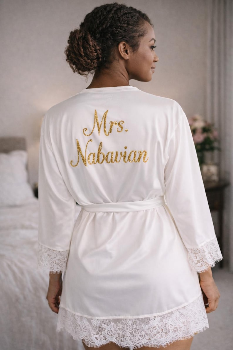 May include: White satin robe with lace trim and a matching belt. The back of the robe features the gold glitter text "Mrs. Nabavian". The robe has long sleeves and is knee-length.
