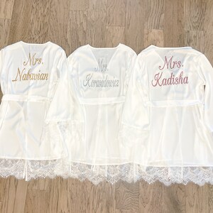 May include: Three white satin robes with lace trim and embroidered names. The robes have wide sleeves and attached belts. The names "Mrs. Nabavian", "Mrs. Korasadowicz", and "Mrs. Kadisha" are embroidered on the back in gold, silver, and pink.