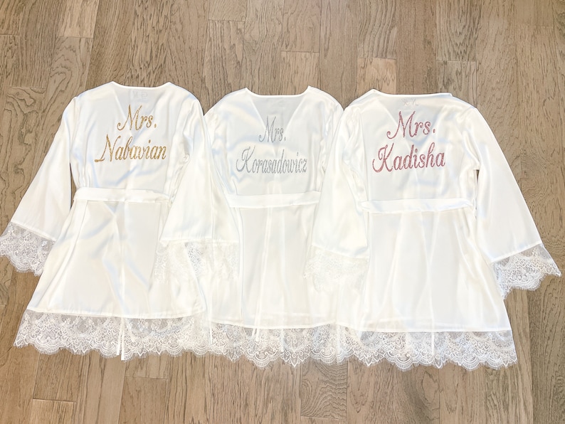 May include: Three white satin robes with lace trim and embroidered names. The robes have wide sleeves and attached belts. The names "Mrs. Nabavian", "Mrs. Korasadowicz", and "Mrs. Kadisha" are embroidered on the back in gold, silver, and pink.