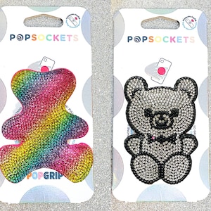 May include: Two PopSockets phone grips featuring a sparkly bear design. One bear is rainbow coloured and the other is silver with black accents. Both have the PopSockets logo and the text "PopGrip".