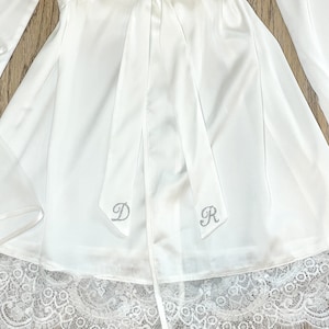 May include: White satin robe with a matching tie, featuring a delicate lace trim at the hem. The robe has long sleeves and is adorned with the silver letters "D" and "R" on the tie. The robe is a bridal or loungewear item.