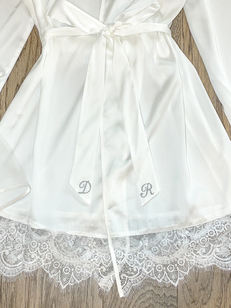 May include: White satin robe with a matching tie, featuring a delicate lace trim at the hem. The robe has long sleeves and is adorned with the silver letters "D" and "R" on the tie. The robe is a bridal or loungewear item.
