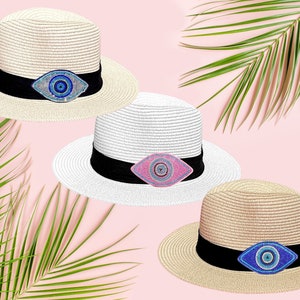 Può includere: Tre cappelli di paglia con ciondoli di occhio turco di diversi colori. I cappelli sono beige, bianco e marrone. I ciondoli di occhio turco sono blu, rosa e blu.
