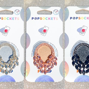 May include: Three PopSockets phone grips with interchangeable tops. The tops are adorned with a cascading design of rhinestones in silver, rose gold, and black.