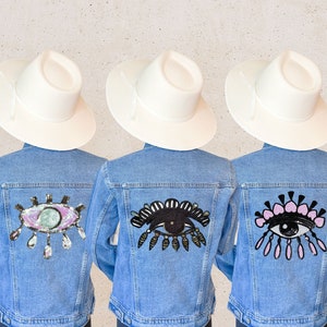 Könnte beinhalten: Drei Jeansjacken mit weißen Cowboyhüten. Jede Jacke hat einen anderen, augenförmigen Patch auf dem Rücken. Die Patches sind aus Pailletten gefertigt und haben unterschiedliche Farben und Designs.