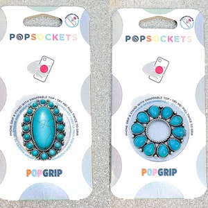 Könnte beinhalten: Zwei türkisfarbene und silberne PopSockets-Handygriffe mit austauschbaren Oberteilen. Die Oberteile zeigen einen türkisfarbenen Stein, der von kleineren türkisfarbenen Steinen umgeben ist. Der Text "PopGrip" ist auf der Unterseite jeder Karte aufgedruckt.