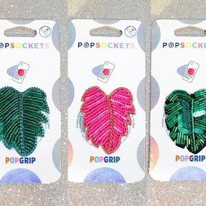 May include: Three phone grips featuring a leaf design. The first grip is green with a blue center, the second is pink with a white center, and the third is green with a white center. Each grip has the text "POPSOCKET" and "POPGRIP" on the packaging.