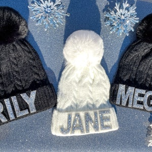 May include: Three knit winter hats with pom poms. The hats are black, white, and gray. Each hat has a name spelled out in silver rhinestones: "Rily", "Jane", and "Megs".
