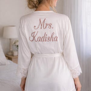 May include: White robe with lace trim and a matching belt. The back of the robe features the text "Mrs. Kadisha" in a glittery rose gold color. The robe has long sleeves and is made of a soft, flowing fabric.