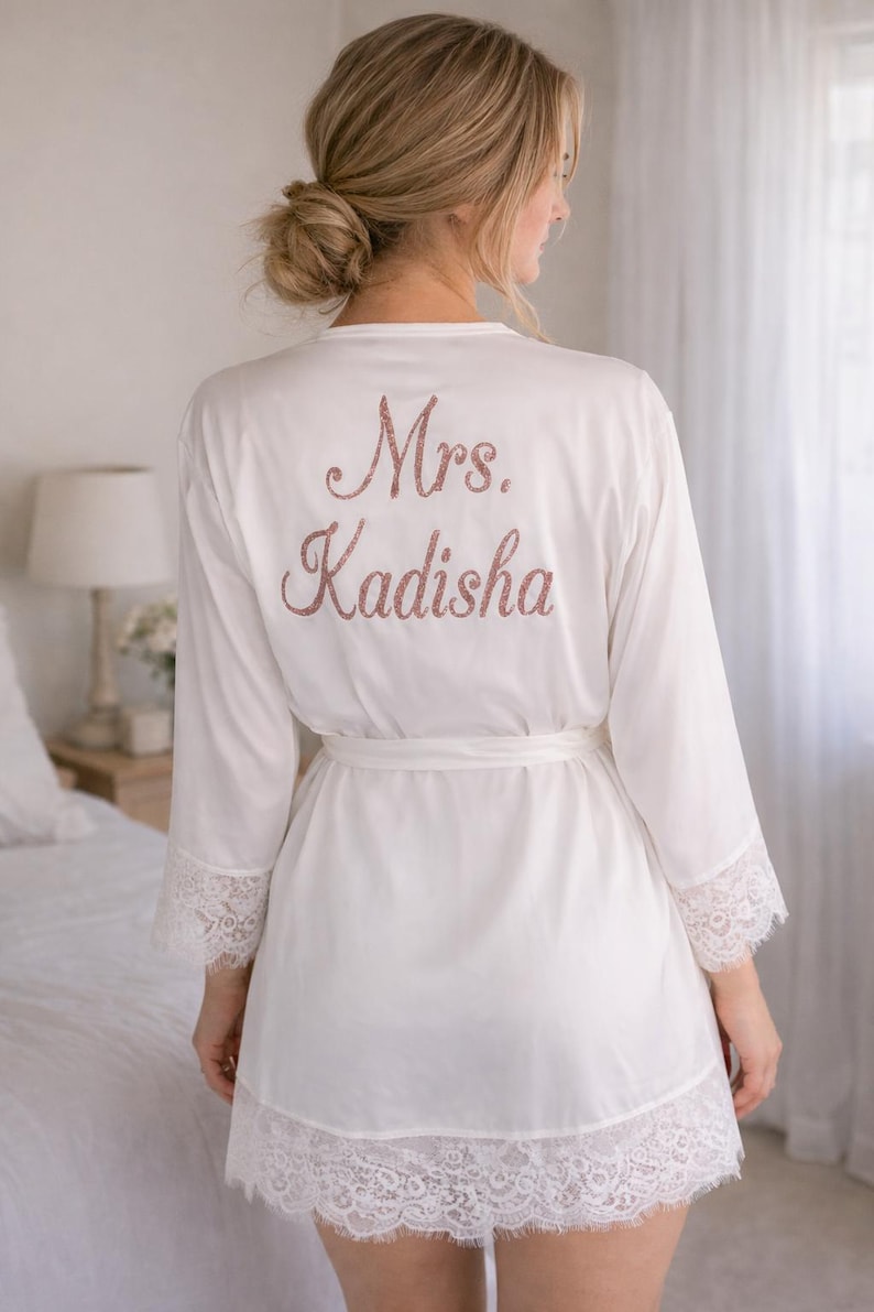 May include: White robe with lace trim and a matching belt. The back of the robe features the text "Mrs. Kadisha" in a glittery rose gold color. The robe has long sleeves and is made of a soft, flowing fabric.