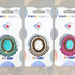 Könnte beinhalten: Drei PopSockets PopGrip Handygriffe mit Perlenmuster. Die Griffe haben ein türkisfarbenes, weißes und rosafarbenes Farbschema mit einem goldenen Akzent. Der Text "PopGrip" ist auf der Unterseite jedes Griffs aufgedruckt.