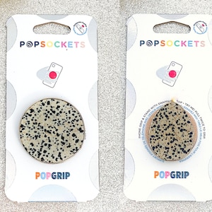 May include: Two packages of PopSockets PopGrip phone grips. The grips are round and have a black and white speckled pattern. The packaging is white with the PopSockets logo and the words "PopGrip" printed on it.