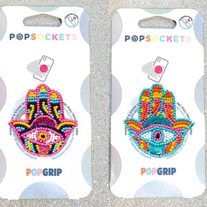 May include: Two PopSockets PopGrip phone grips with a hamsa hand design. One is pink with blue and yellow accents, the other is blue with orange and yellow accents. Both have a beaded design and a white eye.