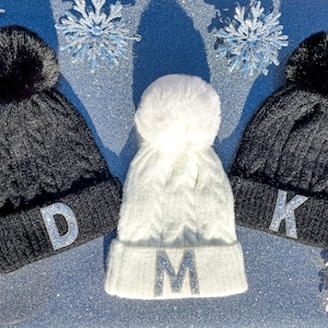 Puede incluir: Tres gorros de invierno con pompones y letras iniciales. Dos gorros negros de punto trenzado con pompones negros y las letras "D" y "K" en pedrería plateada. Un gorro blanco con un pompón blanco y la letra "M".