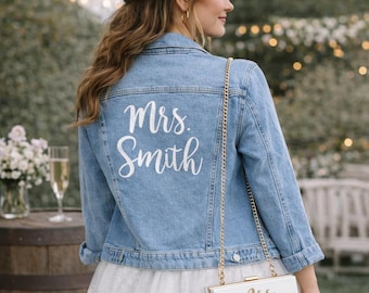 May include: Light blue denim jacket with the embroidered text "Mrs. Smith" on the back. A white clutch with the same text hangs from a gold chain. The jacket is part of a bridal ensemble.