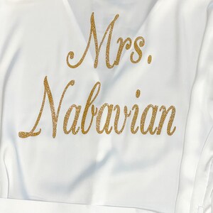 May include: White satin robe with gold glitter lettering that reads "Mrs. Nabavian". The robe has a V-neck and a matching belt. The text is in a cursive font.