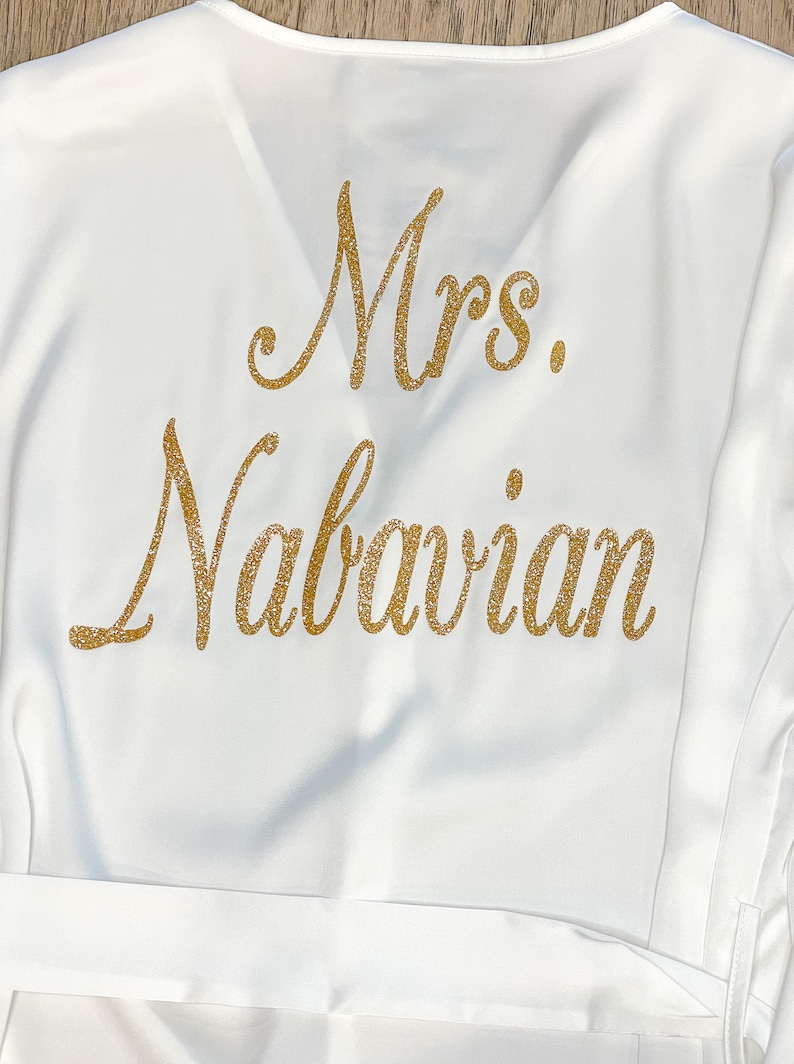 May include: White satin robe with gold glitter lettering that reads "Mrs. Nabavian". The robe has a V-neck and a matching belt. The text is in a cursive font.
