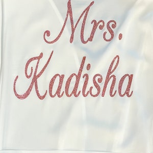 May include: White satin robe with the text "Mrs. Kadisha" in glittery rose-gold script across the back. The robe has a simple, elegant design, perfect for bridal wear or a special occasion.