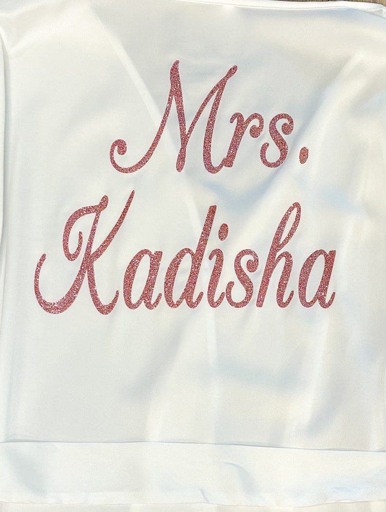 May include: White satin robe with the text "Mrs. Kadisha" in glittery rose-gold script across the back. The robe has a simple, elegant design, perfect for bridal wear or a special occasion.
