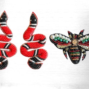 Puede incluir: Un conjunto de tres piezas de joyería de cuentas: dos pendientes de serpiente roja y negra y un broche de abeja negro y verde. La abeja tiene detalles de perlas blancas.