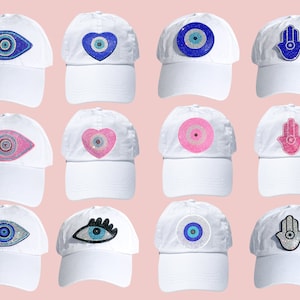 Puede incluir: Doce gorras de béisbol blancas con diferentes diseños de pedrería de colores. Los diseños incluyen un ojo turco azul, un ojo turco rosa, un corazón azul con un ojo turco, un corazón rosa con un ojo turco, un círculo azul con un ojo turco, un círculo rosa con un ojo turco, un ojo turco negro con pestañas, un ojo turco azul con contorno blanco, una mano de hamsa azul, una mano de hamsa rosa y una mano de hamsa azul con contorno blanco.