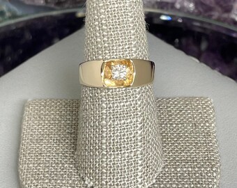 May include: A gold band ring with a single round diamond set in the center. The ring is on a gray fabric ring display stand.