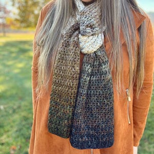 May include: A hand-knitted scarf with a gradient color scheme, transitioning from cream to brown and ending in dark blue. The scarf is made of a textured yarn and is draped around the neck. The background is a blurred outdoor scene.