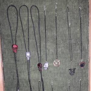 May include: A collection of nine bolo ties with various skull and other decorative pendants. The bolo ties are made of black cord and feature silver, red, and black skull pendants.