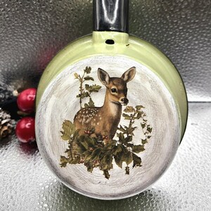 May include: A vintage, light green frying pan with a black handle. The pan features a detailed illustration of a deer surrounded by foliage. The handle is made of metal and has a hole for hanging.