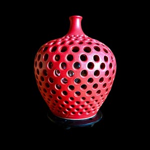May include: A bright red ceramic vase with a rounded body and a narrow neck, featuring a uniform pattern of circular cutouts across its entire surface. The vase is supported by a circular black base, creating a contrast with the vibrant red color.