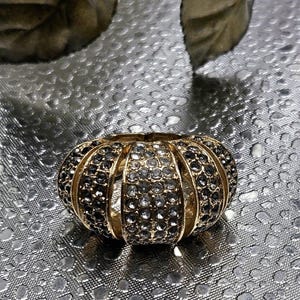 May include: A gold-toned ring with a domed shape, encrusted with alternating rows of black and clear crystals. The ring has a ribbed design and sits on a textured silver surface. The ring's design is reminiscent of a pumpkin.