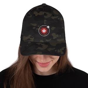 Op de afbeelding: Een zwarte en groene camouflage baseballpet met een rode cirkel en een wit Canadees esdoornbladlogo. Het logo heeft ook een witte cirkel eromheen met een kleine witte ster.