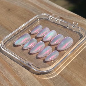 May include: A set of ten almond-shaped press-on nails arranged in a clear plastic storage case. Each nail features a smooth, iridescent finish that shifts between soft white, pale blue, and vibrant pink tones, creating a reflective, pearlescent glow.