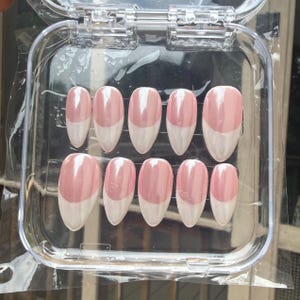 May include: A set of ten almond-shaped press-on nails arranged in a clear plastic storage case. Each nail features a glossy, pearlescent pink base with a distinct white French tip design, creating a smooth, reflective finish across the surface.