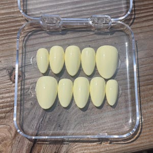 May include: A set of ten almond-shaped press-on nails in a solid pale yellow color with a glossy finish, arranged in two rows inside a clear, square plastic storage container with a hinged lid.
