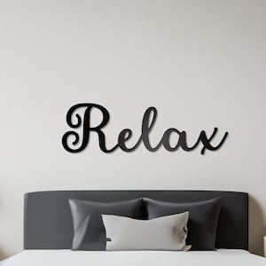 May include: A black metal wall decor with the word "Relax" in a cursive font, above a gray upholstered bed with white bedding and two pillows.