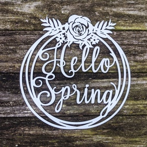 Pu&ograve; includere: Un cartello in metallo bianco con una ghirlanda di fiori e il testo "Hello Spring" in un carattere decorativo.