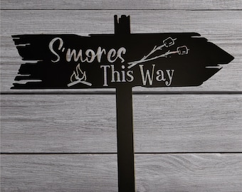Könnte beinhalten: Schwarzes Metallschild mit einer rustikalen Pfeilform, die nach rechts zeigt. Das Schild zeigt "S'mores This Way" mit einer Lagerfeuergrafik und einer Strichmännchenfigur, die zwei Marshmallows hält.