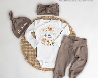May include: A baby outfit set with a white long-sleeved bodysuit featuring a floral wreath with the name "Aubree Grace" in black text. The set also includes a brown headband, a brown beanie with the name "Aubree" in white text, and brown pants.