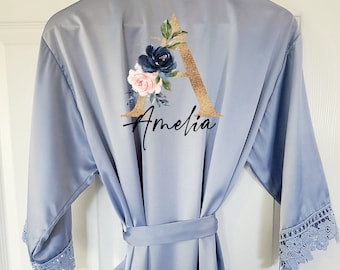 May include: A light blue satin robe with lace trim on the sleeves. The back of the robe features a gold glitter letter "A" with a floral design in navy, pink, and green, and the name "Amelia" in script.