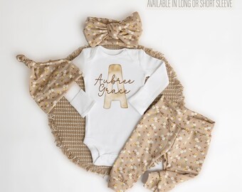 May include: A white baby onesie with long sleeves and the name "Aubree Grace" and a brown letter "A" printed on the front. The onesie is paired with a brown and white floral headband, a brown and white floral hat, and brown and white floral pants.