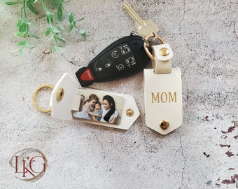 May include: White leather keychain with a photo insert and a separate tag that says "MOM" in gold lettering. The photo insert shows a family of three.