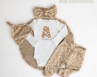 May include: A baby outfit set in a floral print with a white bodysuit featuring the letter A and the name Aubree. The set includes a headband, pants, and a hat.