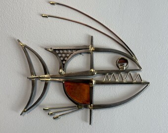 Pode incluir: Uma escultura de metal de um peixe com um design geométrico. O peixe é feito de metal prateado e dourado e tem um olho de vidro vermelho e laranja. A escultura está montada em uma parede branca.