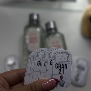 May include: A hand holds a stack of white rectangular stickers with the text "SPICY TAMARINDO" and "GRAN 21". The stickers feature a small image of a baby. Two clear bottles with silver caps are in the background.