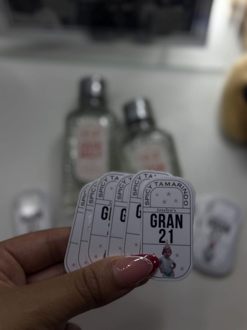 May include: A hand holds a stack of white rectangular stickers with the text "SPICY TAMARINDO" and "GRAN 21". The stickers feature a small image of a baby. Two clear bottles with silver caps are in the background.