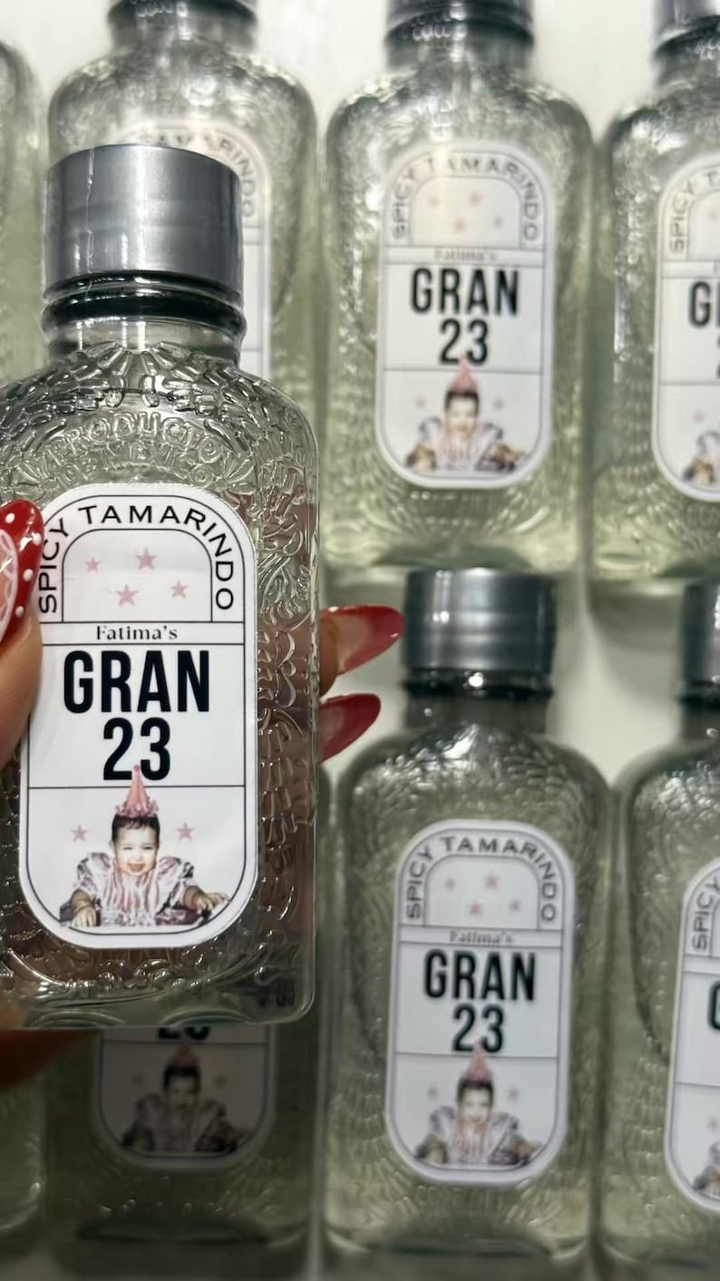 May include: Clear glass bottles with silver caps, labeled "Fatima's GRAN 23". The labels feature a baby in a party hat. The bottles are filled with a clear liquid and are arranged in a grid.